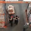 Two warehouse workers in uniform walking next to a pallet jack loaded with boxes in a warehouse aisle.