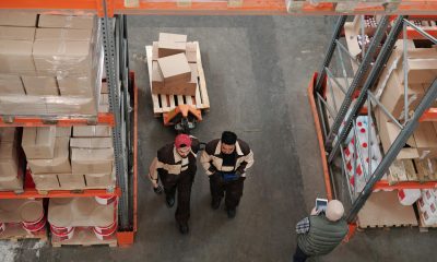 Two warehouse workers in uniform walking next to a pallet jack loaded with boxes in a warehouse aisle.