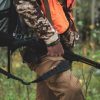 A hunter wearing camouflage and an orange safety vest, holding a bolt-action rifle as they walk through a dense forest.