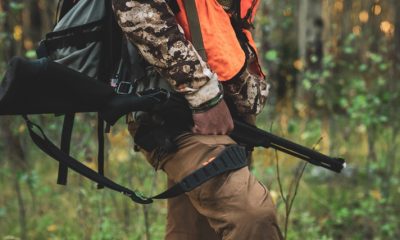 A hunter wearing camouflage and an orange safety vest, holding a bolt-action rifle as they walk through a dense forest.